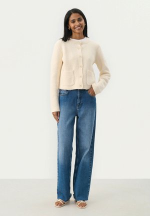 Cardigan crème en tricot avec deux poches à l'avant et un col rond, associé à un jean large bleu clair et des sandales à lanières beiges.