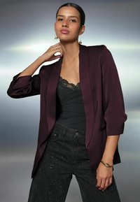 Donna che indossa un blazer bordeaux, un top nero con pizzo, e pantaloni neri con borchie, con orecchini a cerchio dorati e un braccialetto, che posa con sicurezza.