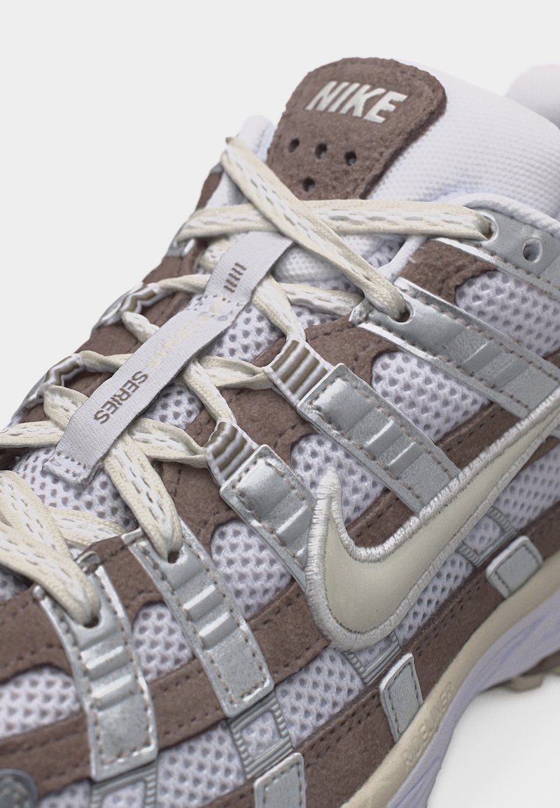 Baskets Nike marron et blanches avec des panneaux en mesh et en suède, des lacets beiges, des accents argentés et le logo Nike swoosh visible sur le côté.