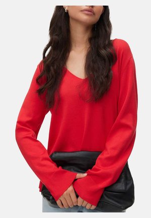 Femeie purtând un pulover roșu cu mâneci lungi și decolteu în V, ținând un clutch mare din piele neagră pe un fundal simplu.