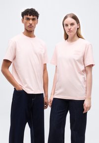 Άνδρας και γυναίκα στέκονται δίπλα-δίπλα φορώντας ροζ ανοιχτόχρωμες κοντομάνικες μπλούζες και σκούρα τζιν παντελόνια με φαρδιές φόρμες, μπροστά σε απλό λευκό φόντο.