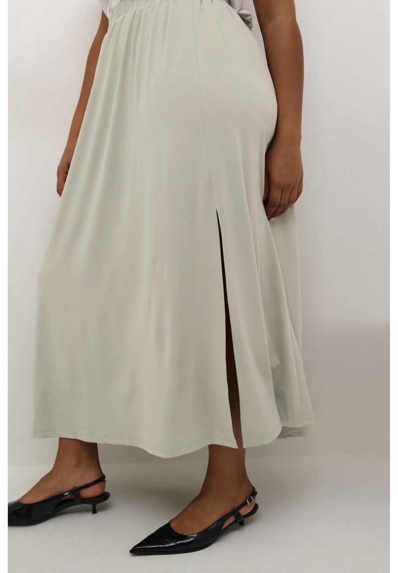 Falda maxi de un gris claro, fluida y con una abertura lateral, hecha de una tela suave. Combinada con zapatos de tacón negro con punta afilada y tiras en el talón.