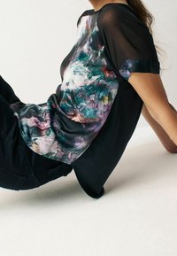 Černý top s krátkým rukávem, který má na přední straně barevný, abstraktní květinový vzor, s hladkou texturou a lehkým materiálem.