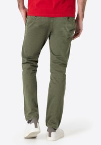 Olivengrüne Chinos mit Slim-Fit, aus Baumwollmaterial, zwei Gesäßtaschen mit Knopfverschluss, getragen mit weißen Sneakern.