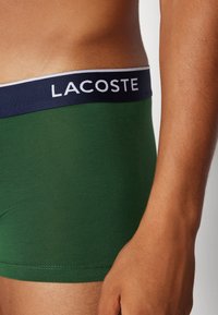 Zelene bokserice s mornarsko plavim pojasom s logotipom "LACOSTE" u bijeloj boji. Tkanina izgleda mekano s glatkom teksturom.