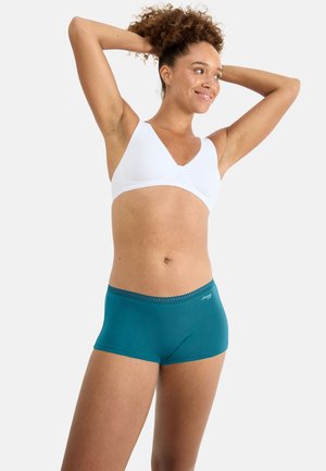 Frau mit lockigem Haar, die einen weißen BH und türkisfarbene Boyshort-Unterwäsche trägt, lächelt mit erhobenen Händen hinter dem Kopf vor weißem Hintergrund.