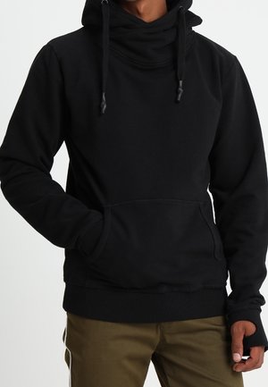 Person iført en sort pullover-hoodie med en frontlomme og snore, kombineret med olivengrønne bukser.