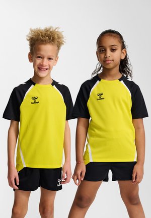 To børn iført matchende gule og sorte sportstrøjer og sorte shorts, der står side om side mod en ensfarvet baggrund.