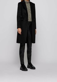 Svart ullkappa med en skräddarsydd design, med en slagkrage och två framfickor, bärs över en mönstrad topp och svarta läderbyxor.