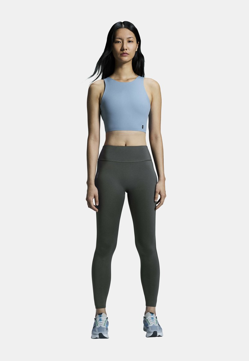 Jasnoniebieski crop top i ciemnozielone legginsy z wysokim stanem o gładkiej fakturze, zestawione z szarymi butami sportowymi z niebieskimi akcentami.
