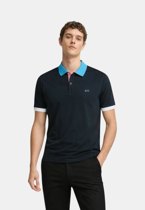 Giovane uomo con indosso una polo nera con colletto blu brillante, patta dei bottoni rosa, polsini bianchi sulle maniche e pantaloni neri, in piedi con una mano in tasca.