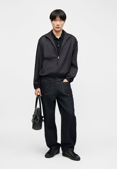 Tânăr purtând o jachetă închisă la culoare cu fermoar, tricou polo negru, blugi largi închiși la culoare, adidași negri, ținând o geantă neagră într-o mână, cealaltă mână în buzunar.