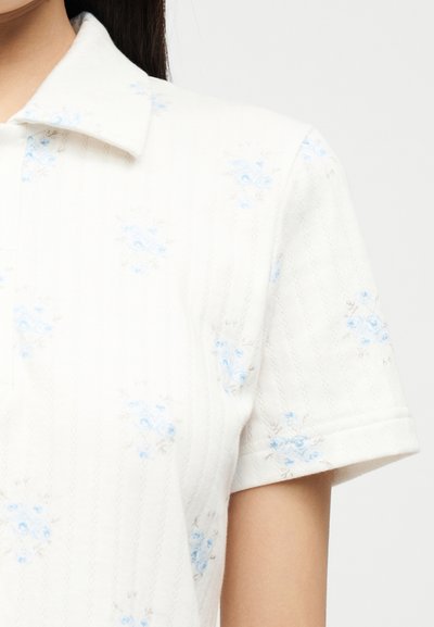 Chemise blanche à manches courtes avec col, ornée d'un motif floral bleu subtil et de lignes verticales texturées, portée par une personne aux longs cheveux bruns.