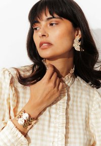 Femme aux cheveux bruns portant un chemisier à carreaux beige, de grandes boucles d'oreilles en or texturé et un bracelet large assorti, posant devant un fond blanc.