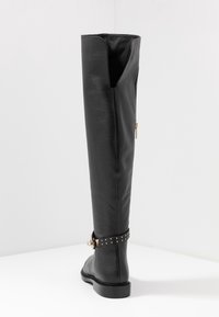 Bota negra de cuero hasta la rodilla con acabado texturizado, accesorios de hardware dorado, cremallera lateral y detalles de correa con tachuelas alrededor del tobillo.
