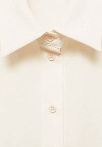 Camicia color crema con colletto classico, sette bottoni e finitura in tessuto testurizzato, che mostra un motivo sottile e materiale morbido.