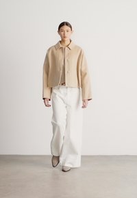 Béžová krátka bunda s golierom a gombíkmi, v kombinácii so širokými bielymi nohavicami. Hladká látka, minimalistický dizajn a neutrálne farby.