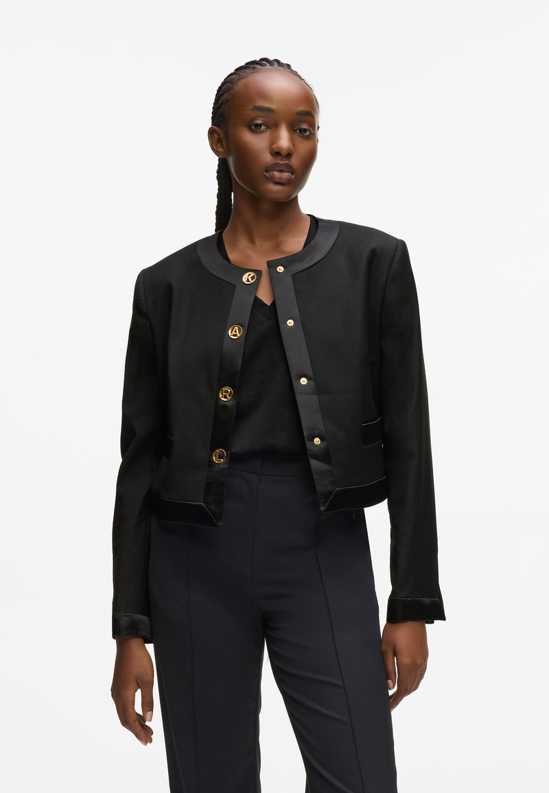 Fekete rövid fazonú dzseki, kerek nyakkivágással, arany gombokkal és selymes részletekkel az ujjakon és az alján. Simaság, szabott sziluett.