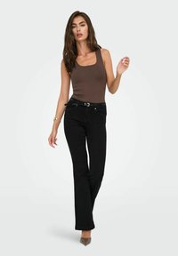 Bruine mouwloze top gecombineerd met zwarte uitlopende jeans voorzien van een riem. Hoge hakken maken de outfit compleet. Eenvoudige achtergrond.