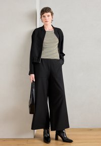 Veste noire raccourcie sur un haut rayé vert et blanc, associée à un pantalon large noir et des bottes à talons noirs brillantes.