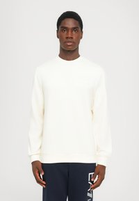 Joven vestido con un sudadera blanca de manga larga sencilla y pantalones azul marino con texto gráfico blanco en la pierna izquierda, de pie frente a un fondo blanco.