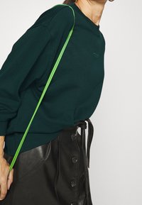Persona che indossa un maglione verde scuro e una gonna di pelle nera con bottoni, con un cinturino di borsa verde brillante appoggiato sulla spalla.