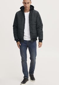 Giacca nera imbottita con cappuccio, zip frontale e tasche laterali, abbinata a una camicia bianca e jeans blu. Indossata con sneakers nere.