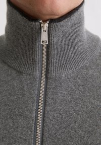 Grauer Strickpullover mit hohem Kragen, ausgestattet mit einem silbernen Reißverschluss und geripptem Textur an Kragen und Körper.