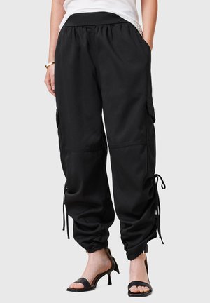 Femme portant un pantalon cargo jogger noir avec poches latérales et cordons ajustables, associé à des sandales à talons noires et un haut blanc.