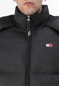 Svart dunjakke med høy krage, frontglidelås og stripet snor. Har en liten "Tommy Jeans" logo på venstre bryst.