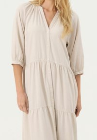 Robe boutonnée couleur crème avec un col en V, des manches trois-quarts et une jupe à volants froncés. Tissu doux et léger avec une texture lisse.