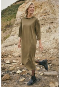 Robe à capuche vert olive avec manches trois-quarts, texture douce et fentes latérales, associée à des bottines noires sur un terrain rocheux.