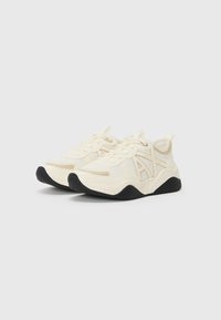 Cremfarbene Sportschuhe mit strukturiertem Obermaterial, ausgestattet mit einer kontrastierenden schwarzen Sohle und dezenten Logo-Details an den Seiten. Rundliche Zehenform.