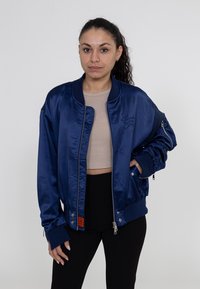 Marineblaue Satin-Bomberjacke mit gerippten Bündchen und Saum, Reißverschlusstaschen und Logo-Stickerei auf der Brust. Glatte Textur insgesamt.