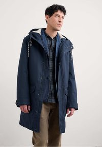 Námornícka parka s kapucňou s umelou kožušinou, prednými vreckami a zapínaním na gombíky na manžetách. Nosená cez kostkovanú košeľu, má uvoľnený strih po kolená.