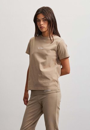 Giovane donna con capelli lunghi indossa una maglietta beige e pantaloni abbinati, in piedi con un braccio dietro la schiena su sfondo neutro.