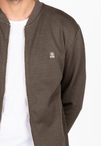 Veste aviateur marron en tissu texturé, avec un col côtelé et une fermeture éclair sur le devant. Elle possède un petit logo brodé sur la poitrine gauche.
