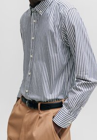 Randig skjorta med knappar i marinblått och vitt, tillverkad av lätt tyg, med klassisk krage och långa ärmar, kombinerad med beige byxor.