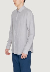 Camicia grigia chiara a righe con colletto, maniche lunghe e bottoni blu, abbinata a pantaloni blu scuro. Tessuto con una texture sottile visibile.