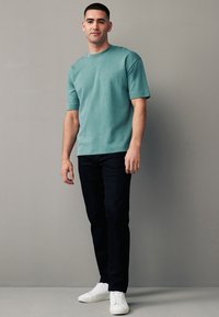 Helles türkisfarbenes T-Shirt mit kurzen Ärmeln aus weichem Stoff, kombiniert mit dunkelblauen Jeans und weißen Sneakern, vor einem neutralen Hintergrund stehend.