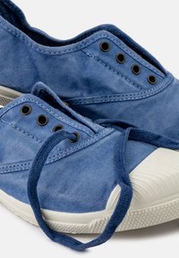 Kék vászon sportcipők fekete fűzőlyukakkal, fehér gumitalppal és texturált orral. A fűzők lazán megkötve pihennek a cipőn.