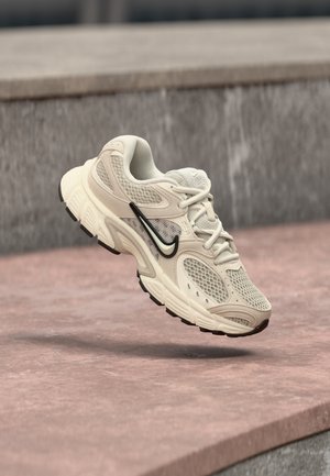 Beige and white athletic sneaker with mesh and suede details, black Nike swoosh, captured mid-air above a pink concrete surface.