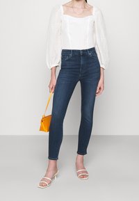 Blusa blanca texturizada con mangas abullonadas, combinada con jeans ajustados de cintura alta en azul oscuro. Los accesorios incluyen un pequeño bolso naranja y sandalias blancas.