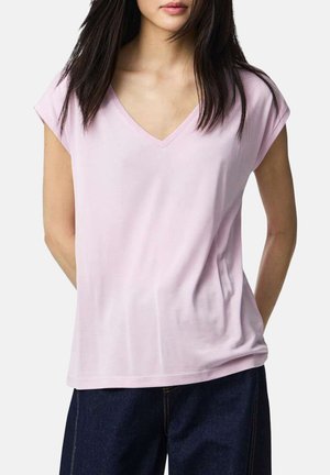 Femme portant un haut rose pâle à manches courtes et col en V, associé à un pantalon en denim bleu foncé, debout avec les mains derrière le dos.