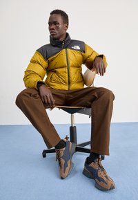 Geel en zwarte pufferjack, bruine wijde broek en bruine/blauwe sneakers. Het model zit op een houten stoel in een blauwe kamer.