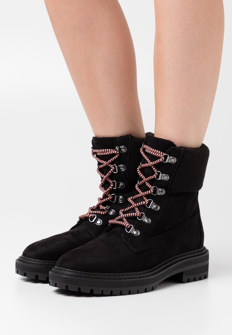 Botines de ante negros con suelas gruesas, cordones rosa y negros, usados sobre piernas desnudas contra un fondo blanco.