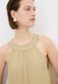 Beiges ärmelloses Kleid mit hohem Kragen, das sanfte Falten und offene Seiten hat. Große goldene Ohrringe mit einer pinken Mitte und strukturiertem Rand.