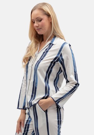 Donna con lunghi capelli biondi che indossa un pigiama bianco con strisce verticali blu, in piedi con una mano in tasca e guarda verso il basso.