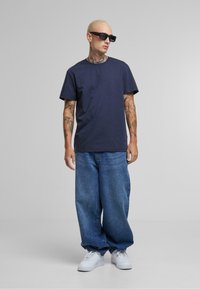 Tricou din bumbac bleumarin, asortat cu blugi denim lejeri albaștri și adidași albi. Ochelarii de soare adaugă un accent îndrăzneț. Design simplu și casual.