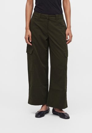 Femme portant un pantalon cargo vert foncé à jambes larges avec poches à rabat et des ballerines noires sur fond blanc.